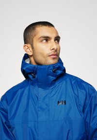 Chaqueta impermeable azul real con un alto cuello, capucha ajustable y un logo negro liso en el pecho. Material suave y brillante.