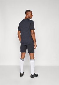 Zwart sportshirt met textuurdetails, gecombineerd met zwarte shorts. Witte sokken met drie zwarte strepen en zwarte voetbalschoenen.