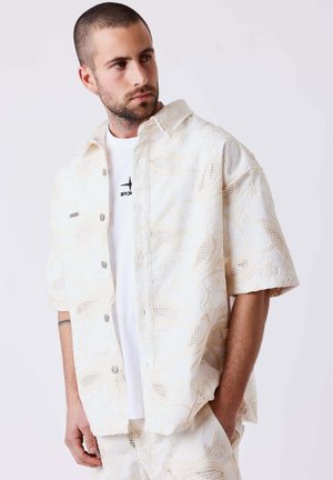 Homme aux cheveux courts et à la barbe porte une chemise blanche à manches courtes à motif floral et un short assorti, regardant sur le côté avec une main dans la poche.