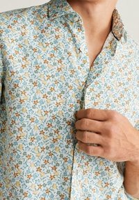 Eine Person, die den Knopf eines hellblauen Hemdes mit kleinem orangefarbenem und gelbem Blumenmuster schließt, wobei Hals- und Handdetails zu sehen sind.