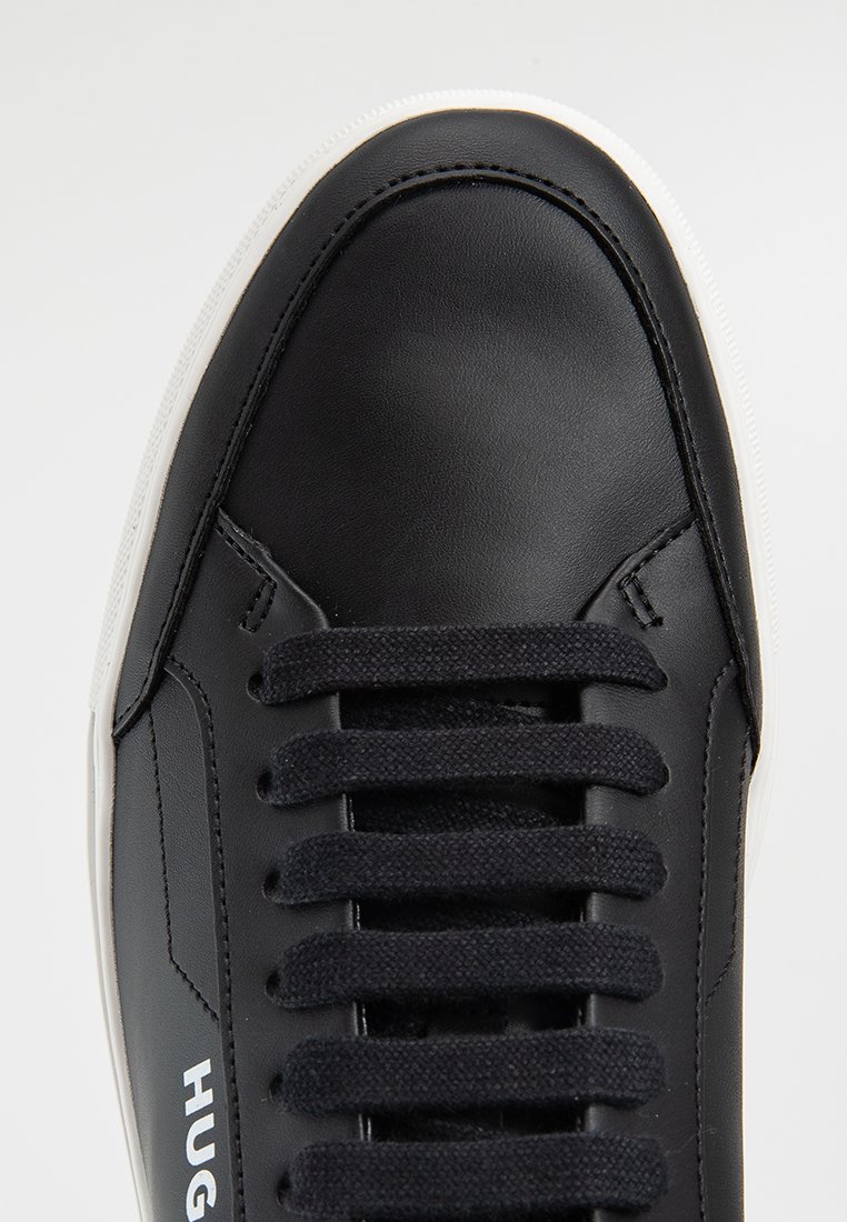 Schwarze Leder-Sneaker mit schwarzen Schnürsenkeln, weißer Sohle und dem Schriftzug "HUGO" in weiß an der Seite, von oben betrachtet.