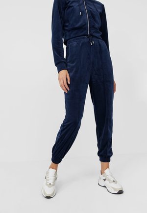 Person iført marineblå velour joggingbukser med elastiske manchetter og hvide atletiksneakers, stående på en ensfarvet lys baggrund.