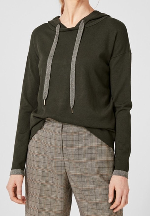 Personne portant un pull à capuche vert foncé avec des cordons gris et un pantalon à carreaux gris, debout devant un fond uni.