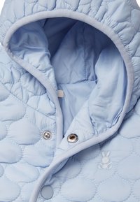 Světle modrá prošívaná bunda s kapucí, která má kulatý vzor, patenty a knoflíkové zapínání, a malé vyšívané detaily králíčka.