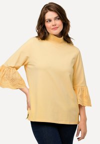 Top giallo a maniche corte con collo alto e maniche svasate, con ricamo ad occhio di pernice. Tessuto morbido, vestibilità rilassata e spacchi laterali.