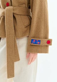 Hnedý vlnený kabát s opaskom, červenými gombíkmi a modrou náplasťou na manžete rukáva, nosený cez svetlé nohavice, zobrazená ruka a dlaň.