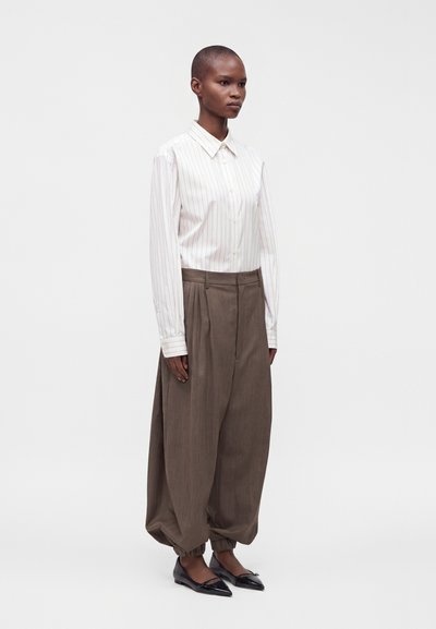 Personne portant une chemise blanche à fines rayures avec boutons, un pantalon marron ample avec des poignets froncés, et des ballerines noires pointues, debout les bras le long du corps.