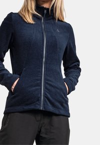 Mörkblå, zip-up fleecejacka med hög krage, utrustad med två sidofickor och en mjuk textur, kombinerad med svarta byxor.