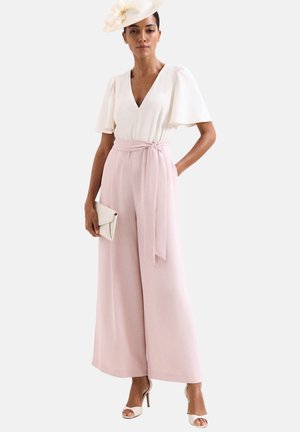 Vrouw draagt een witte V-hals blouse, lichtroze wijde broek met riem, witte open-toe hakken, houdt een witte clutch vast en draagt een witte bloemenhoed.