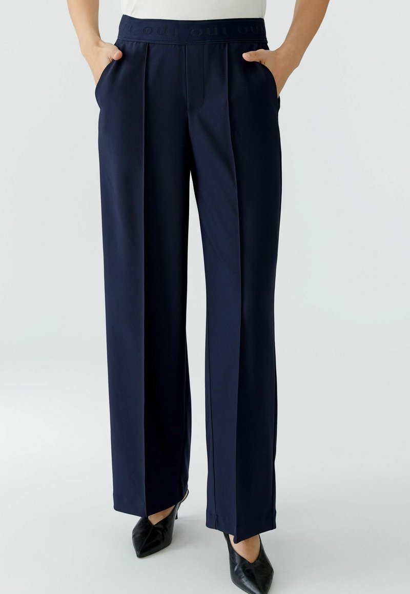 Navy wide-leg trousers featuring a textured fabric, pleated front, and a tonal waistband with logo detail. Side pockets included.