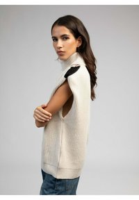 Pull col roulé en crème avec des épaules découpées et des patches noirs. Texture côtelée et coupe ample, associé à un jean bleu.