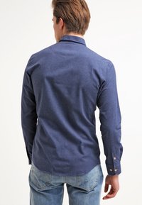 Námořnická modrá košile s dlouhými rukávy a knoflíky, vyrobená z texturované látky. Má zakřivený lem a manžety na dva knoflíky pro nastavitelnou velikost.