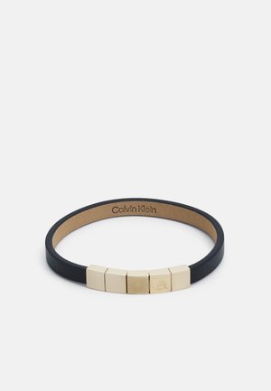Svart läderarmband med fem guld geometriska detaljer och "CK"-gravyr. Slät textur med en minimalistisk design.