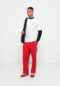 White short-sleeve t-shirt with black trim and logo, black long-sleeve undershirt, red track pants with white stripes, and red shoes.