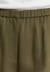 Olijfgroene, lichtgewicht stofbroek met een elastische tailleband, een zijzakdetail en een gladde textuur. Close-up weergave van het taillegebied.