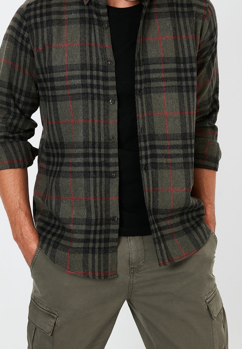 Man i en grön och svart rutig skjorta med knappar över svart t-shirt, med uppvikta ärmar och gröna cargobyxor, händerna i fickorna.