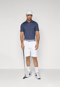 Marineblå polo med røde detaljer og prikkete mønster, kombinert med hvite shorts og hvite golf sko. Holder en golfkølle og har på seg en hvit caps.