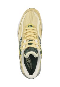 Zapatilla de malla y tela amarilla con acentos verdes, cordones redondos, cuello acolchado y suela blanca y gris. Presenta la marca "New Balance".