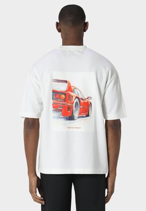 Homme portant un t-shirt blanc avec illustration d'une voiture de sport rouge vue de dos, debout devant un fond uni.