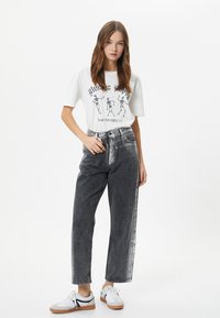 Hvid grafisk t-shirt med sort skeletprint, parret med højtaljede, løstsiddende mørkegrå jeans og hvide sneakers med brune såler.