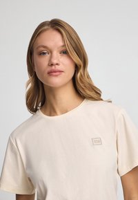 Camiseta de algodón beige de manga corta y escote redondo. Presenta un pequeño parche con un logotipo rectangular en el pecho. Textura suave.