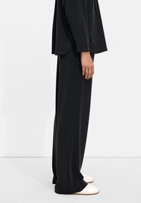 Black, loose-fitting top with long sleeves and rounded hem paired with matching wide-leg pants. White sneakers visible at the bottom.
