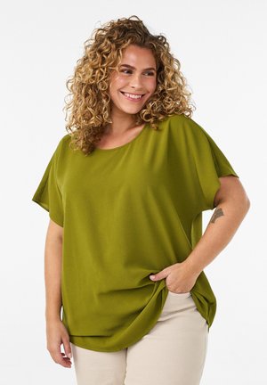 Kvinde med krøllet blondt hår, smilende, iført en oversize grøn kortærmet top og lysebeige bukser, den ene hånd i lommen.