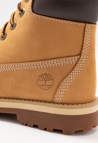 Botte en cuir tan avec un col en cuir marron, des coutures blanches et un logo d'arbre embossé sur le côté. Semelle en caoutchouc avec traction.