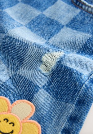 Denimtyget har ett rutigt mönster, med ett fransigt hål och en gul blomsterbroderi nära botten, vilket ger en färgkontrast.