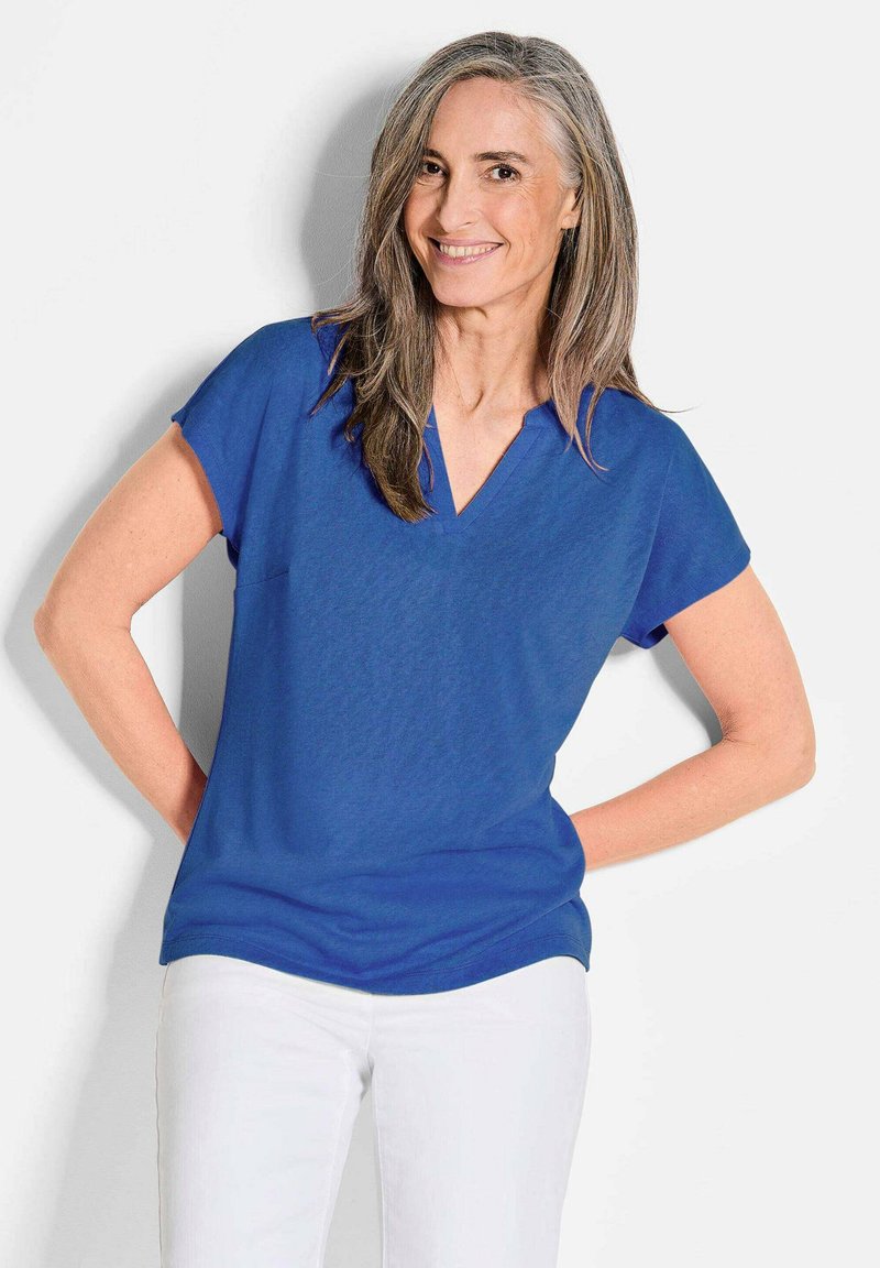 Blauwe top met V-hals en korte mouwen, gemaakt van een gladde stof. Gepaard met witte broek, poseert het model tegen een effen achtergrond.