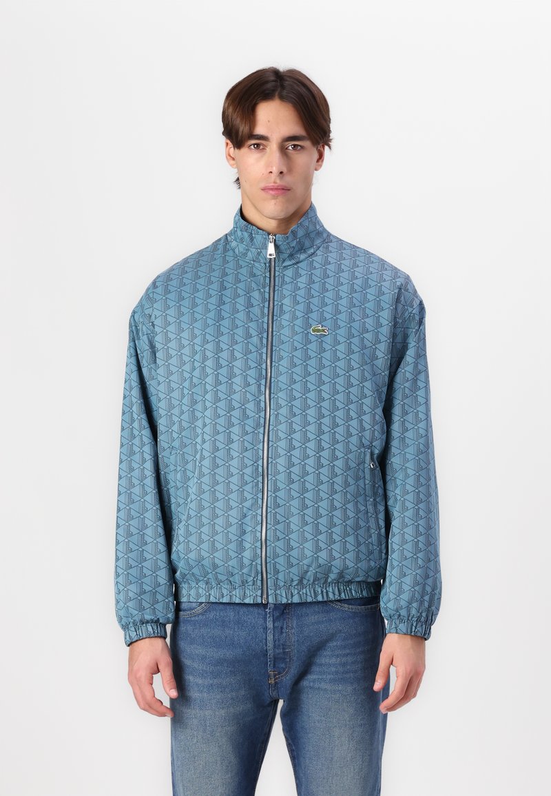 Light blue patterned bomber jacket with a high collar, full zipper, and elastic cuffs. Features a small logo on the left chest area.