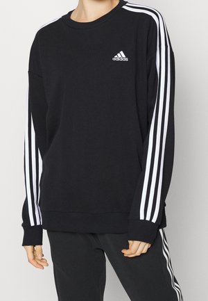 Czarna bluza i spodnie Adidas z białymi potrójnymi paskami na rękawach i nogawkach, noszone przez osobę z rękami swobodnie opuszczonymi wzdłuż ciała.