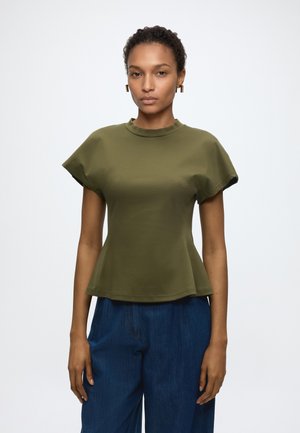 Mujer con top peplum verde oliva de manga corta y pantalones azules de pierna ancha, de pie sobre un fondo blanco liso.