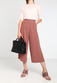 Kvinna iklädd en blushfärgad topp med rynkade ärmar, rosafärgade vida beskarna byxor, rosa klacksandaler, och bär en svart läderhandväska.