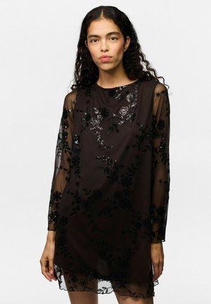 Mujer con cabello largo y rizado viste un vestido negro translúcido con encaje floral y detalles de lentejuelas, de pie frente a un fondo claro y liso.