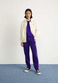 Cilvēks stāv iekštelpās uz zila paklāja, ģērbies purpura Adidas treniņtērpā, krēmkrāsas žaketē un melnās kedās ar baltām auklām.