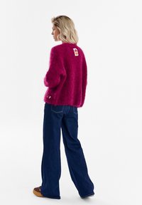 Kobieta o krótkich blond włosach, ubrana w miękki magentowy sweter i szerokie ciemnoniebieskie dżinsy, stojąca bokiem na białym tle.