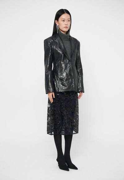 Blazer de couro preto com um acabamento texturizado sobre um suéter gola alta cinza. Abaixo, uma saia azul marinha transparente com bordados florais pretos e meias-calças pretas.