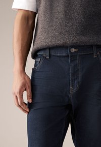 Tummansiniset farkut, joissa on muotoiltu malli, kontrastiväriset oranssit tikkaat, etutaskut ja napitus vyötäröllä.