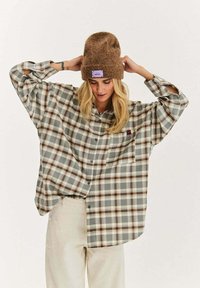 Kobieta poprawiająca brązową dzianinową czapkę, ubrana w oversize'ową szarą i brązową koszulę w kratę oraz kremowe spodnie, stojąca na jednolitym tle.