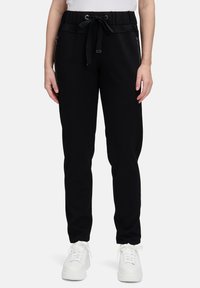 Osoba w czarnych spodniach dresowych z zaszewkami bocznymi i białymi sneakersami, stojąca na tle jednolitym.