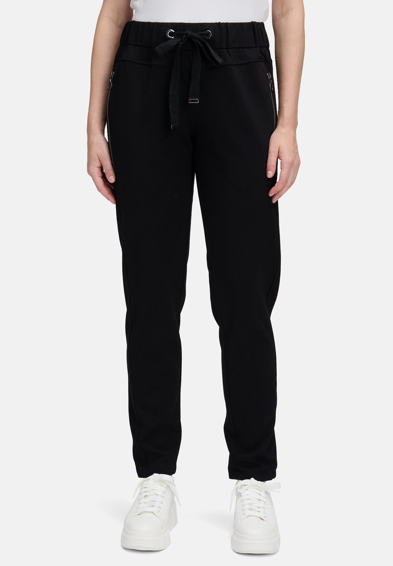 Osoba w czarnych spodniach dresowych z zaszewkami bocznymi i białymi sneakersami, stojąca na tle jednolitym.