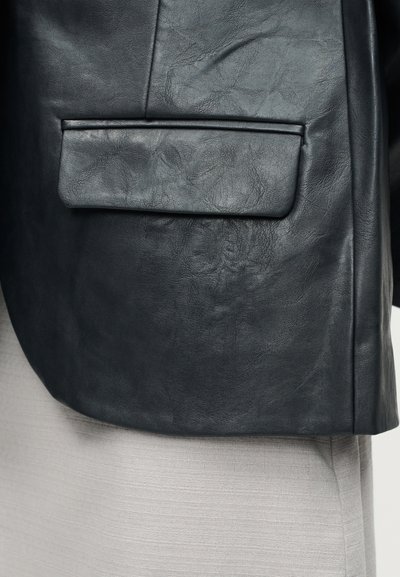 Τσέπη από μαύρο δερμάτινο υλικό με υφή και καπάκι, πάνω σε ανοιχτό γκρι ύφασμα ή ρούχο από κάτω.