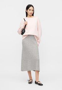 Γυναίκα που φοράει ανοιχτό ροζ φαρδύ πουλόβερ, γκρι midi φούστα με πλέξη καλωδίου, μαύρη τσάντα χιαστί και μαύρα παπούτσια Mary Jane, στέκεται μπροστά σε λευκό φόντο.