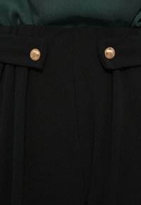 Black trousers featuring a textured fabric, pleats, and gold-tone buttons at the waistband, providing a fitted and structured look.