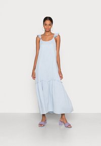 Šviesiai mėlyna maksi suknelė su raukšlėtais petnešomis ir daugiasluoksniu dizainu, pagaminta iš minkštos, tekstūruotos medžiagos. Suderinta su šviesiai purpuriniais blokiniu kulnu sandalais.