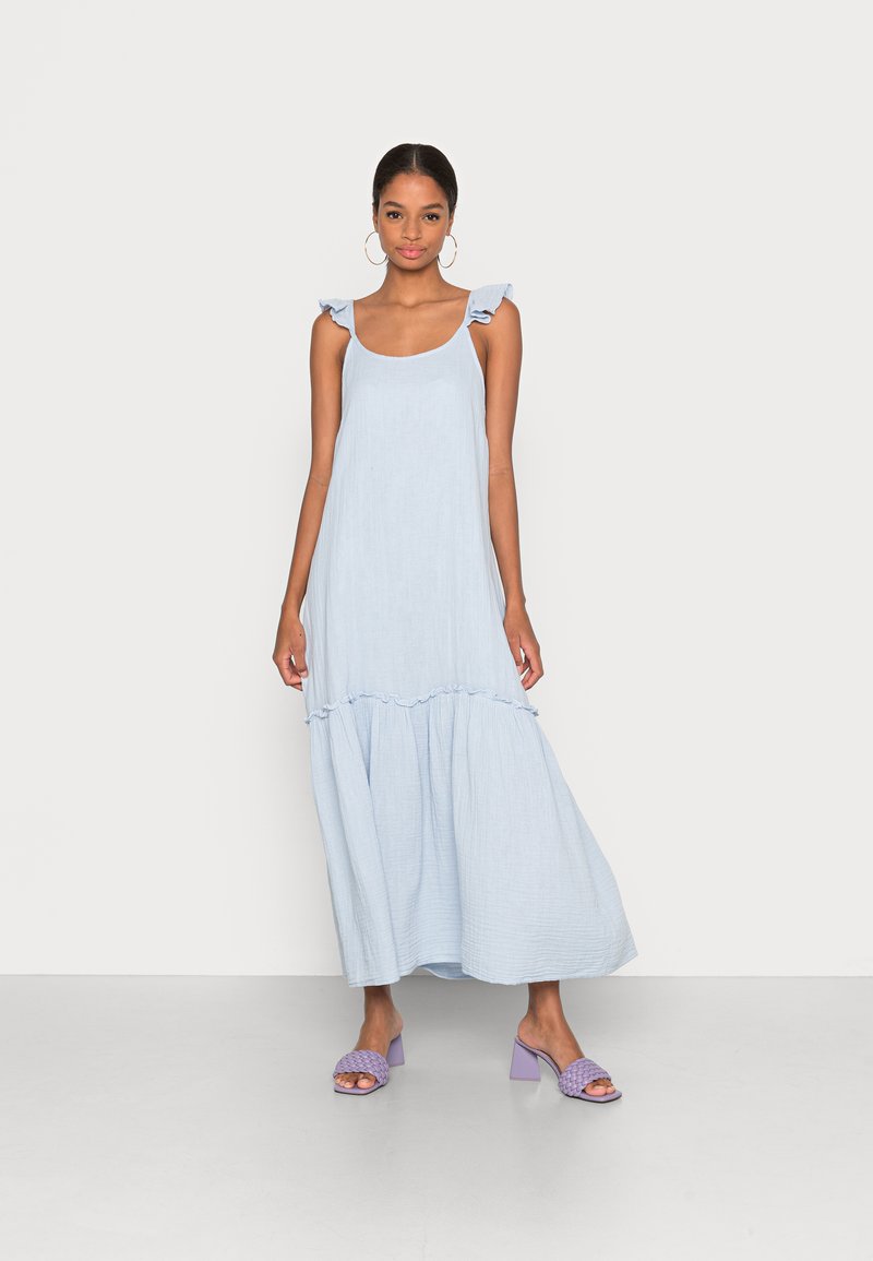 Šviesiai mėlyna maksi suknelė su raukšlėtais petnešomis ir daugiasluoksniu dizainu, pagaminta iš minkštos, tekstūruotos medžiagos. Suderinta su šviesiai purpuriniais blokiniu kulnu sandalais.