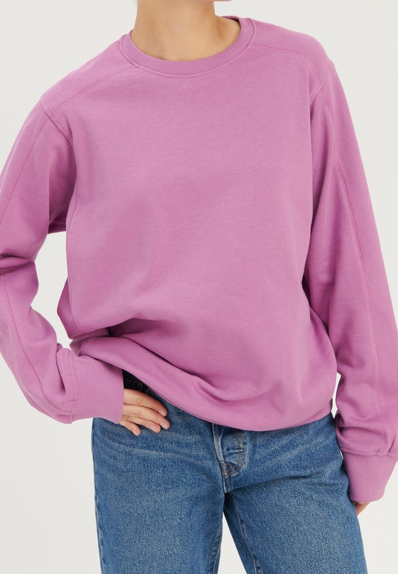 Personne portant un sweat-shirt mauve à manches longues et un jean bleu, avec une main posée sur la hanche, devant un fond uni.