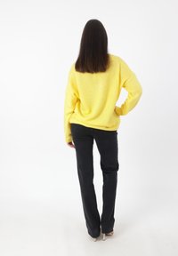 Übergroßer gelber Strickpullover mit gerippten Bündchen, kombiniert mit schwarzen gerade geschnittenen Hosen und hellen Schuhen, von hinten betrachtet.