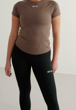 Kobieta ubrana w brązowy dopasowany top z krótkim rękawem i czarne legginsy, oba z białym logo "aimn", stojąca na jasnym jednolitym tle.